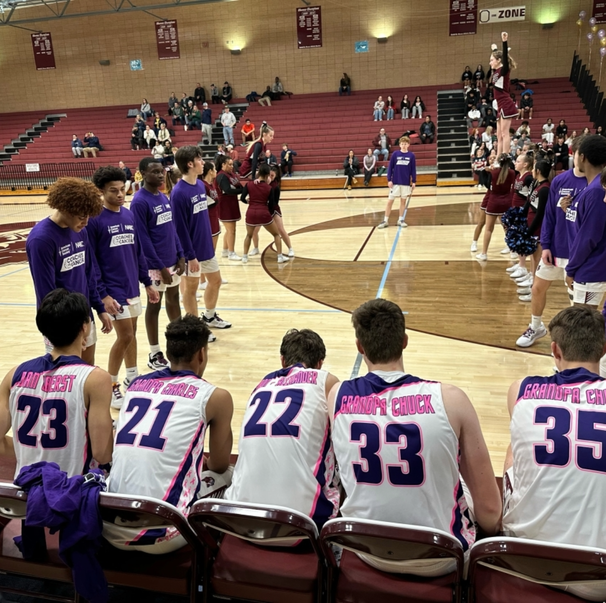 A basketball game in progress at an indoor gymnasium with players and cheerleaders, with spectators seated on bleachers in the background.