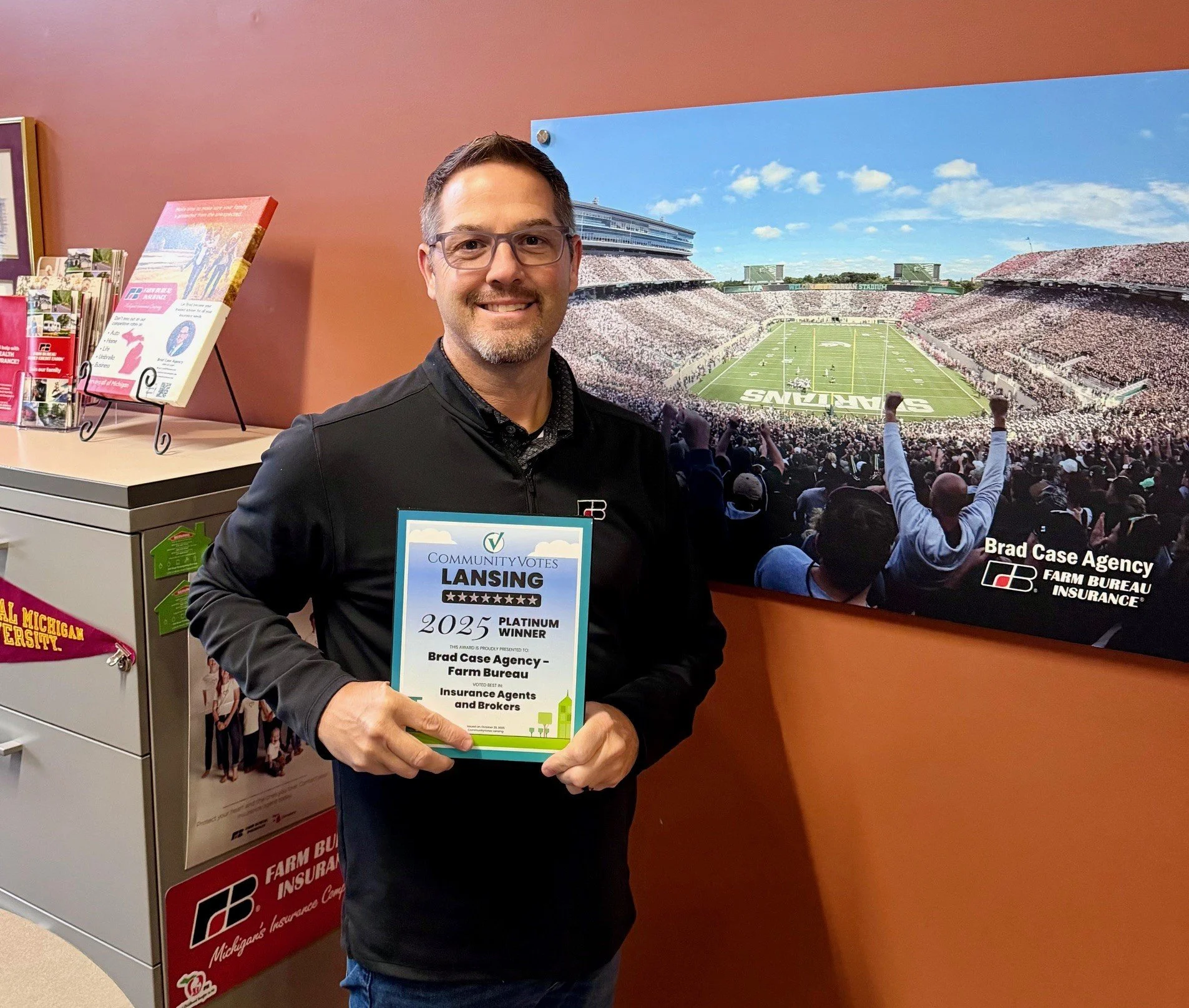 A man in glasses and a black jacket smiling and holding a plaque that says '2023 Platinum Winner'. The background features a large photo of a football stadium filled with spectators and a few brochures on a counter with Michigan State University branding.