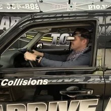 A man driving a black car, wearing sunglasses and a baseball cap, inside a parking lot.