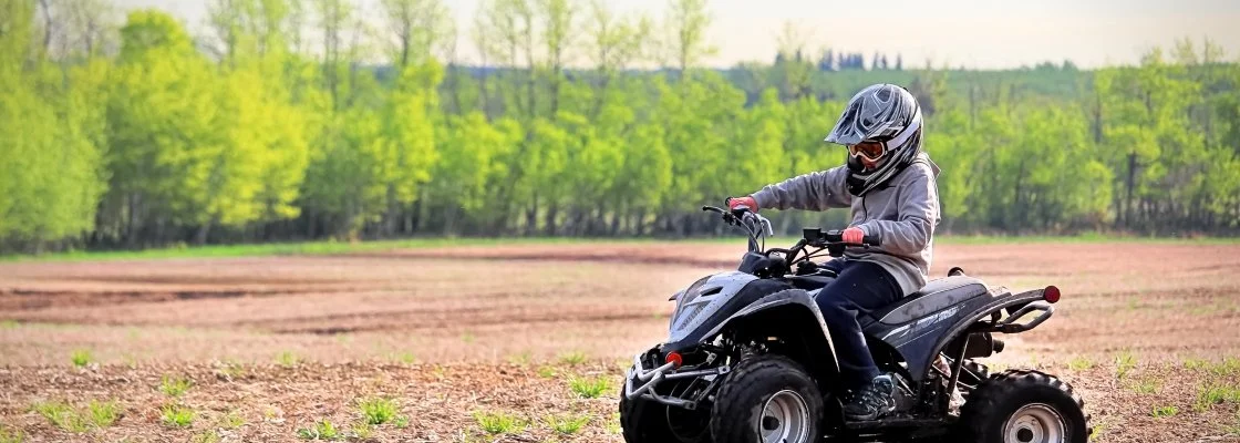 A child riding an all-terrain vehicle (ATV) on a dirt field, wearing a helmet and safety gear, with green trees in the background.