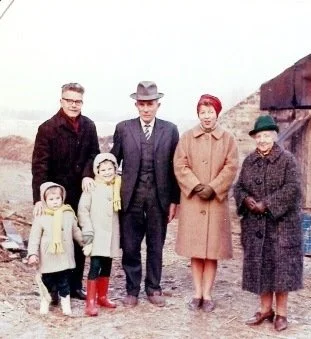 A family of six standing outdoors in front of a hillside. Two young children wearing yellow coats and red boots standing next to an adult woman dressed in a tan coat and hat. Two adult men and an older woman are also present, all dressed warmly for cool weather.