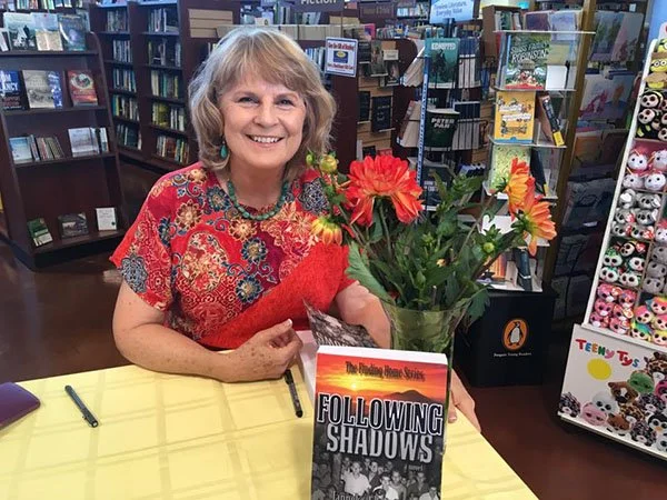 A smiling woman with gray hair holding a bouquet of large orange flowers at a book signing event in a bookstore.