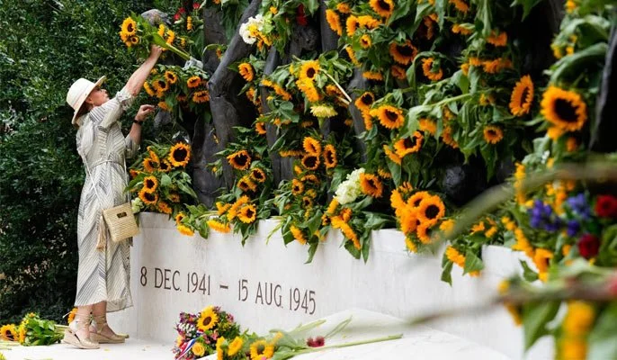 A woman placing flowers at a war memorial decorated with sunflowers, with dates 8 Dec 1941 - 15 Aug 1945 engraved on the white wall.