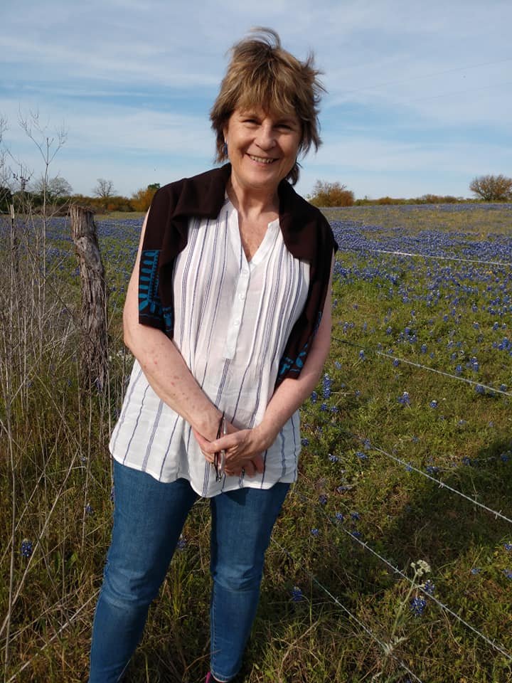 Smiling woman standing in a field of blue flowers, wearing a white striped blouse and jeans, with a jacket draped over her shoulders, during daytime.