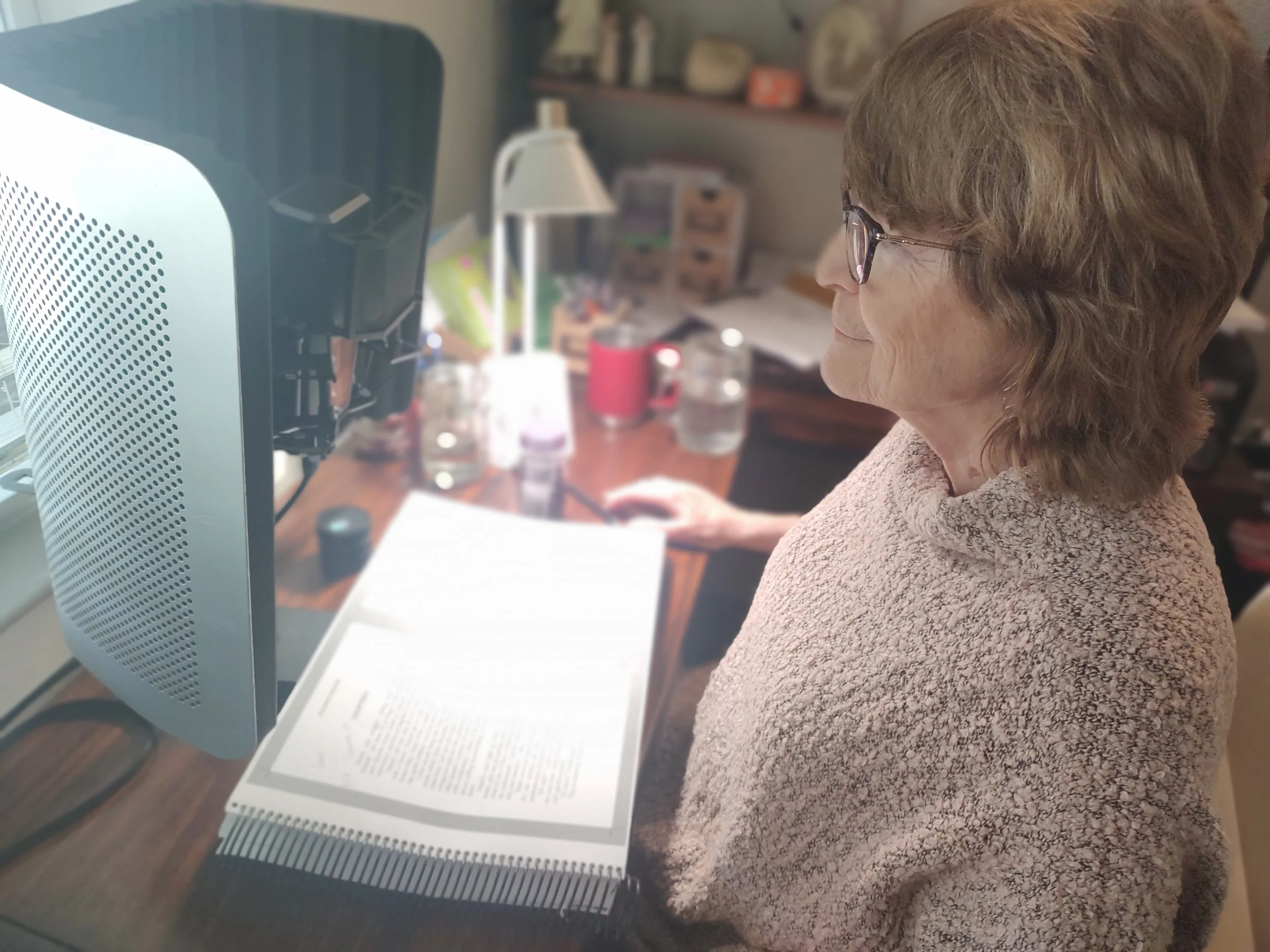 A woman with glasses and curly hair working on a computer at a cluttered desk, with an open book in front of her.