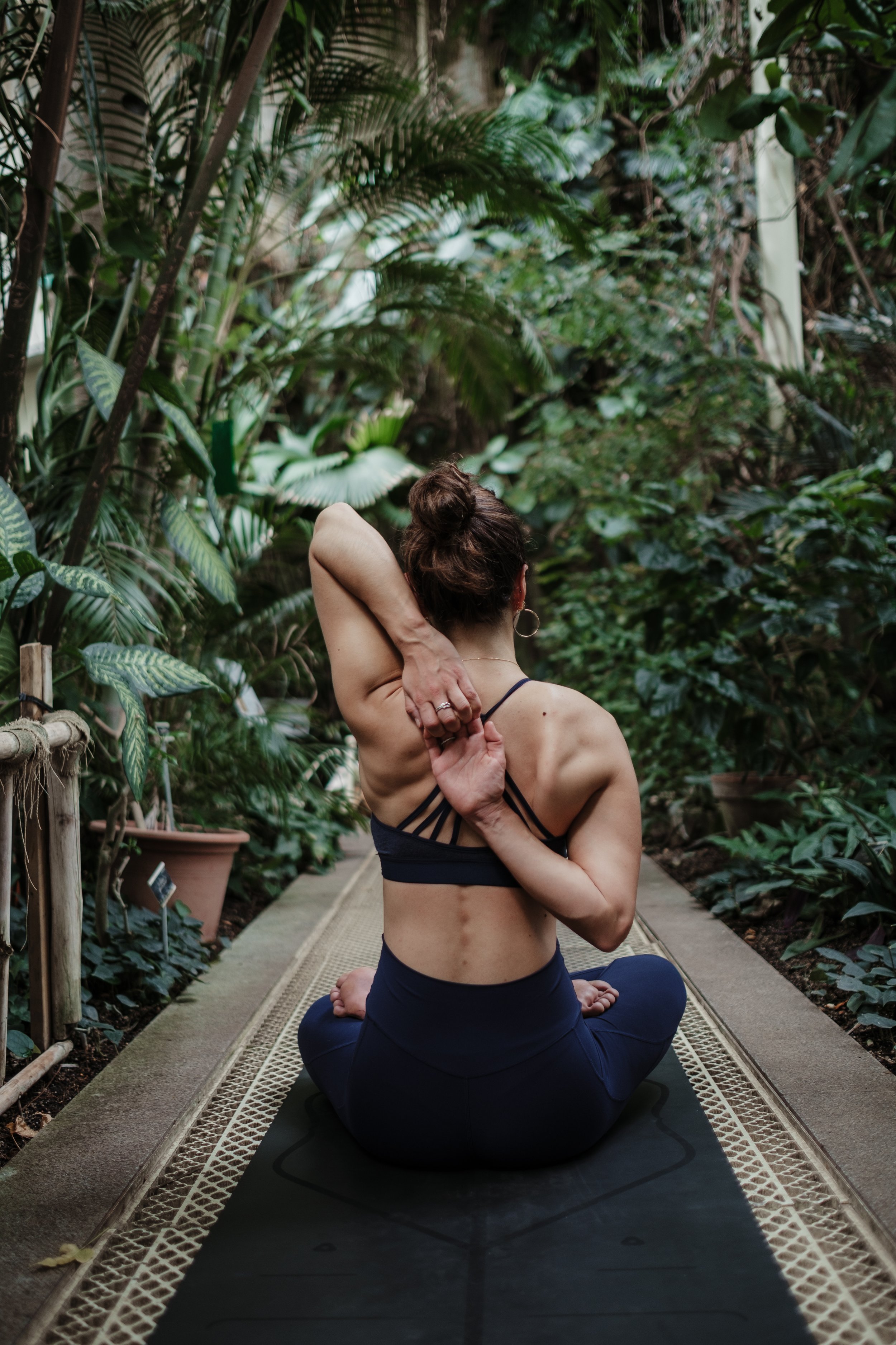 A woman practicing yoga or meditating on a mat in a lush, green conservatory or greenhouse filled with various plants.