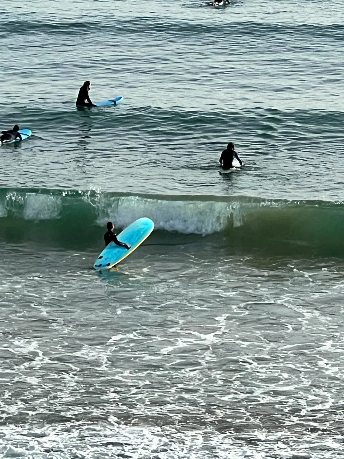 People surfing in the ocean, with one person sitting on a surfboard near the shore and others paddling out to surf further away.