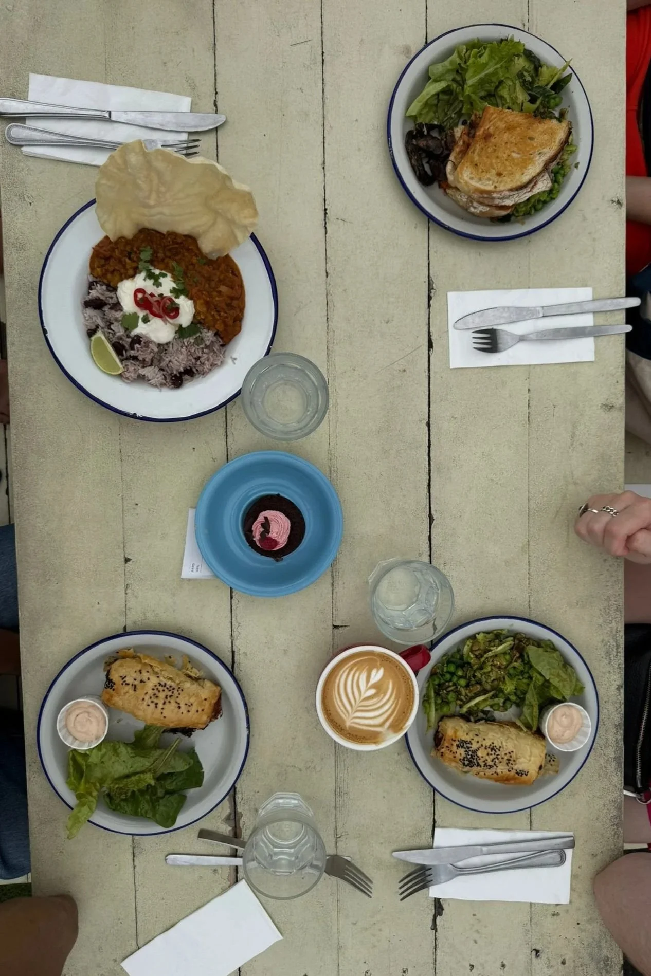 L-shaped table with three people dining, featuring plates of various foods including sandwiches, salads, a piece of cake, and beverages like coffee and water.