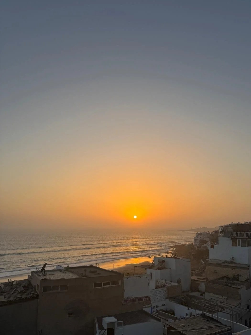 Sunset over the ocean with a beach and buildings in the foreground.