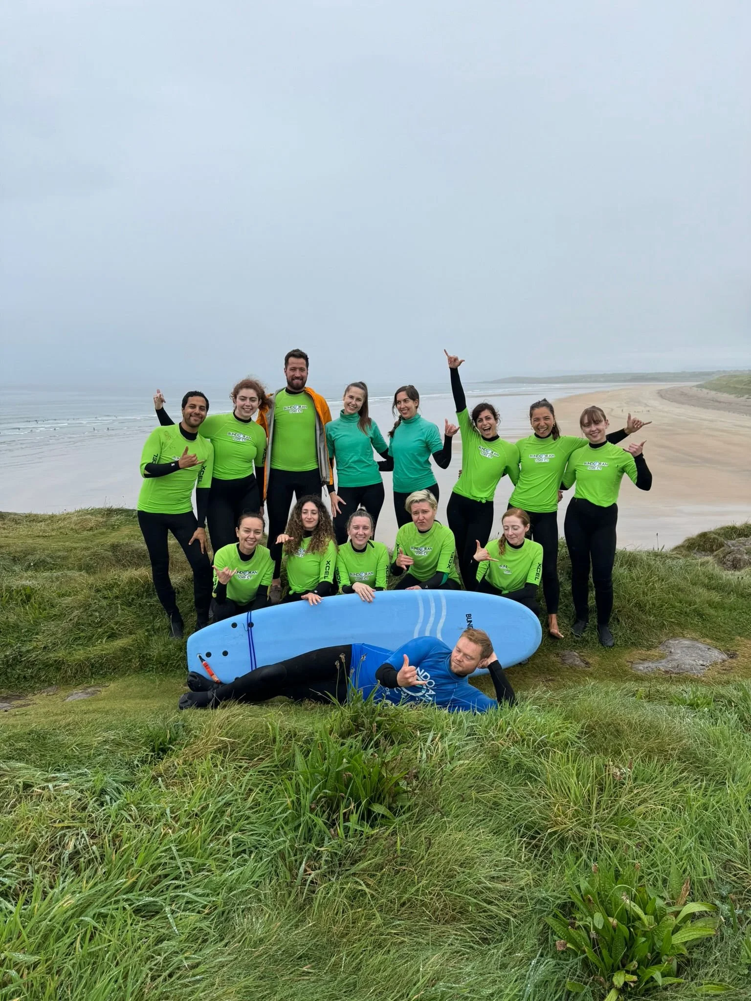 Group of mostly women and one man in green and blue wetsuits and life jackets standing and lying on a grassy hill near a beach, with an overcast sky in the background. They are smiling and making 'shaka' hand gestures.