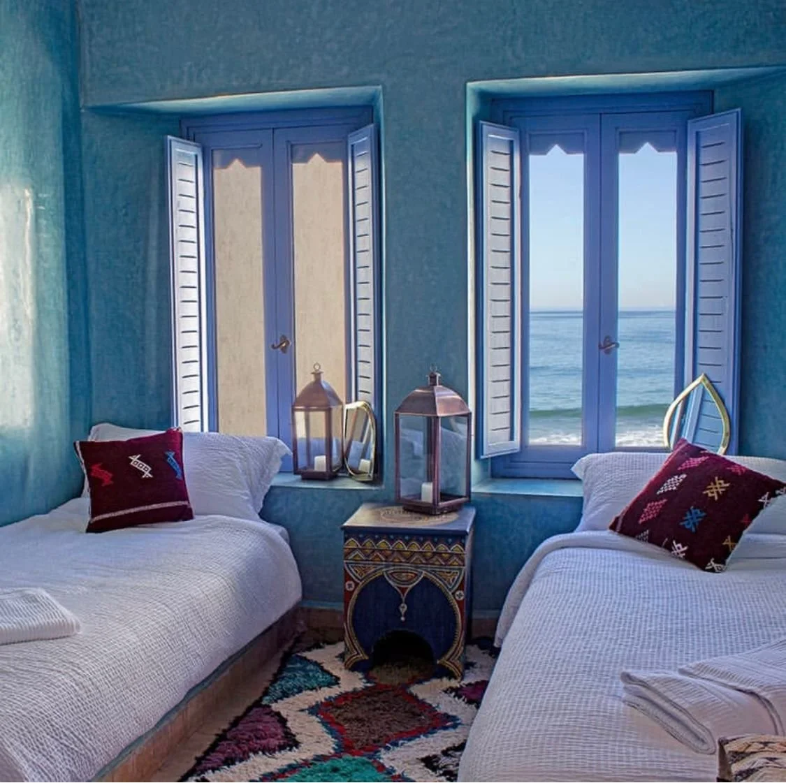 Bedroom with two beds, each with a decorative pillow, and a small colorful table with two lanterns on it. The room has two open windows showing a view of the ocean.