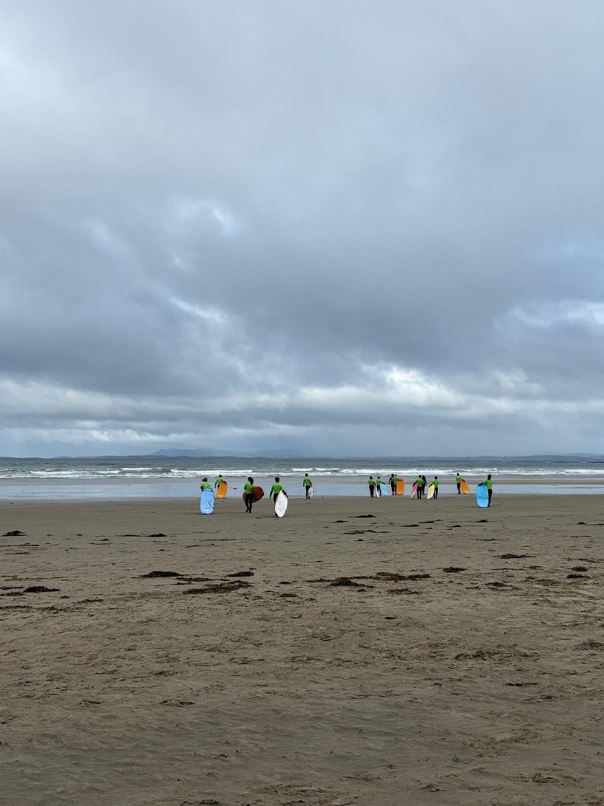 A group of surfers in neon green shirts, holding surfboards of various colors, walking towards the ocean on a cloudy beach.