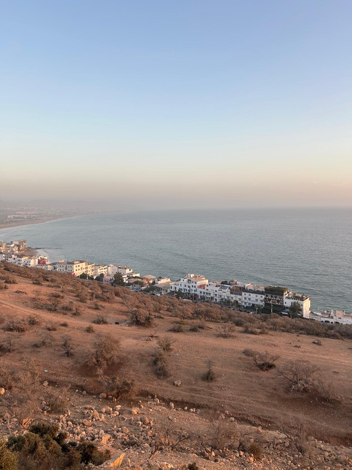 A coastal hillside with dry, sparse vegetation and rocks, overlooking a beach and a row of white buildings along the shoreline under a clear sky.