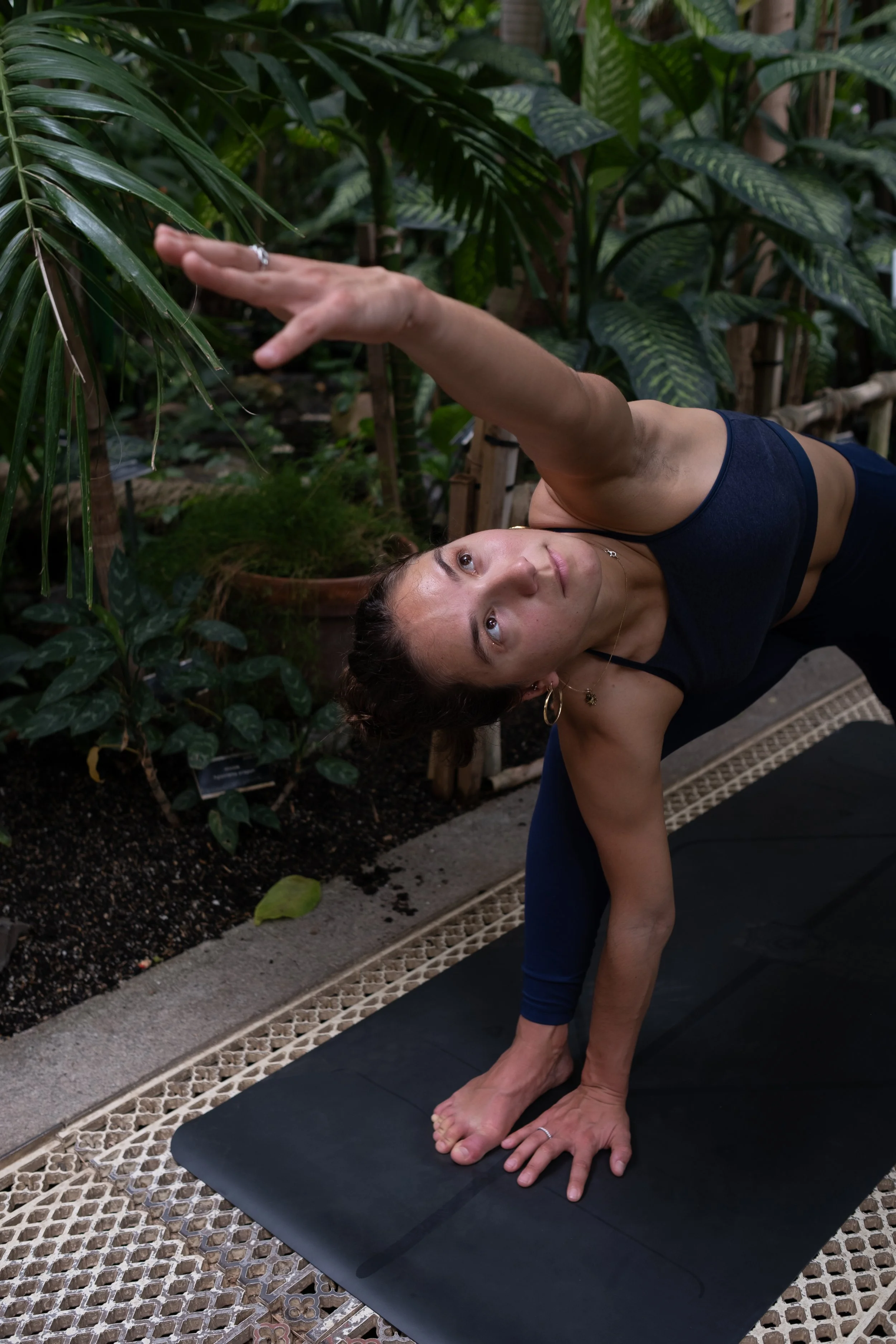 A woman doing yoga in a tropical greenhouse, performing a side plank pose with one arm extended towards the ceiling, on a black yoga mat amidst lush green plants.