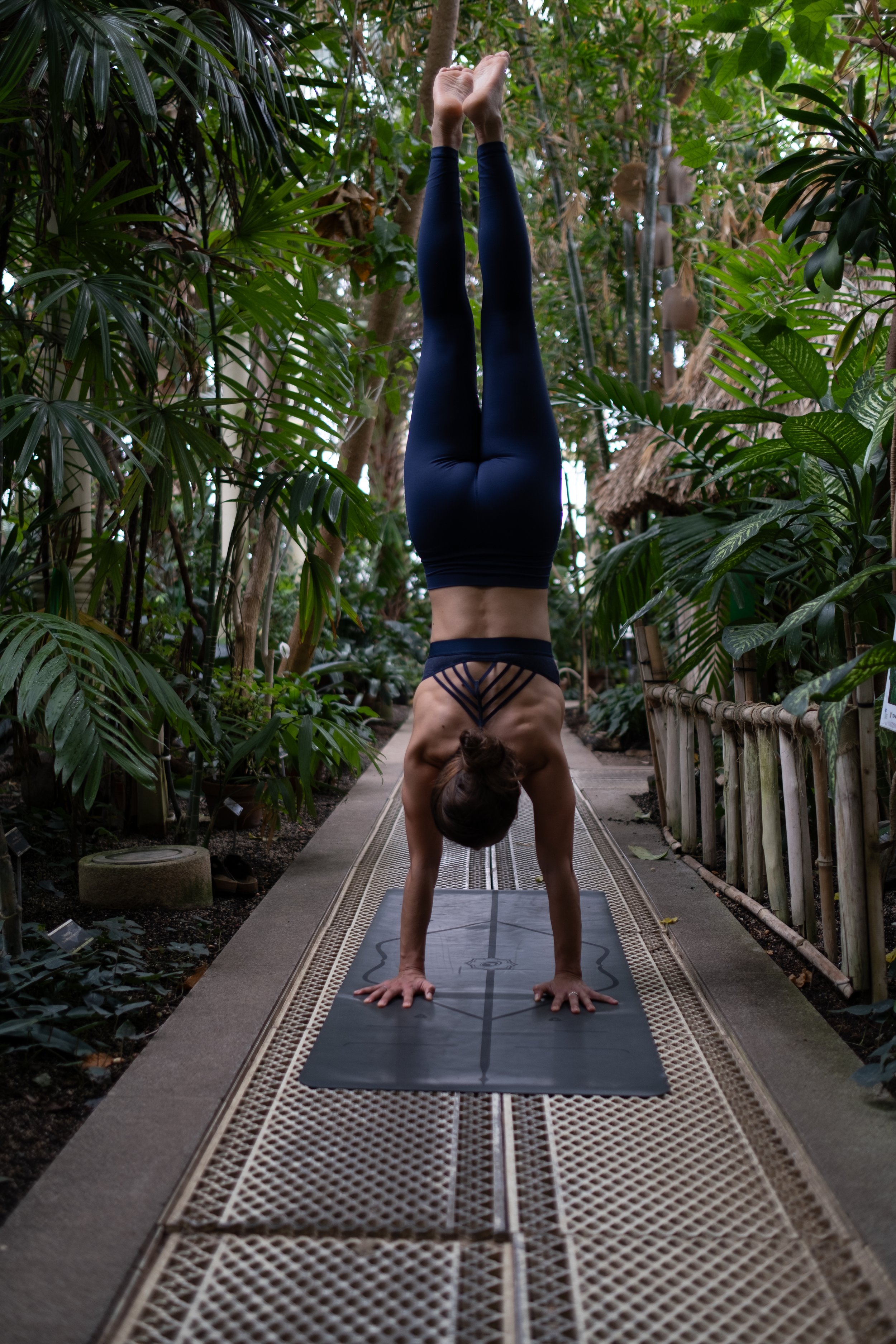 A woman practicing a handstand in a lush indoor garden or greenhouse, standing on a yoga mat on a narrow walkway surrounded by green plants.