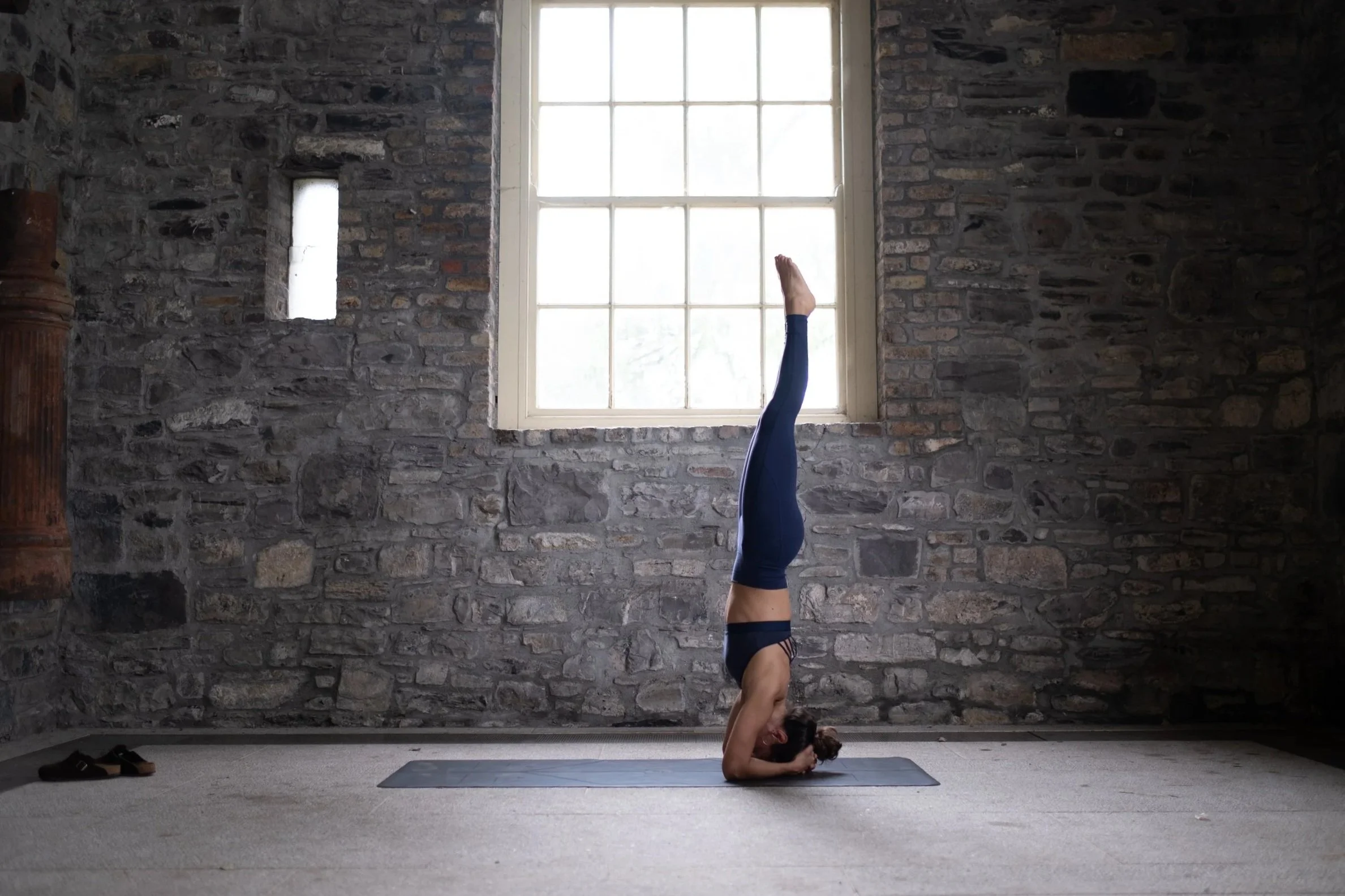 Woman practicing yoga in a headstand pose inside a room with stone walls and a large window.