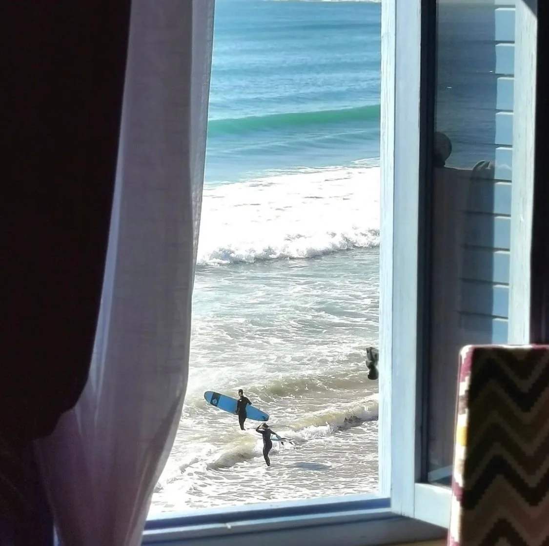 View of people at the beach surfing seen through a window with curtains. Two individuals carrying surfboards are near the shoreline.