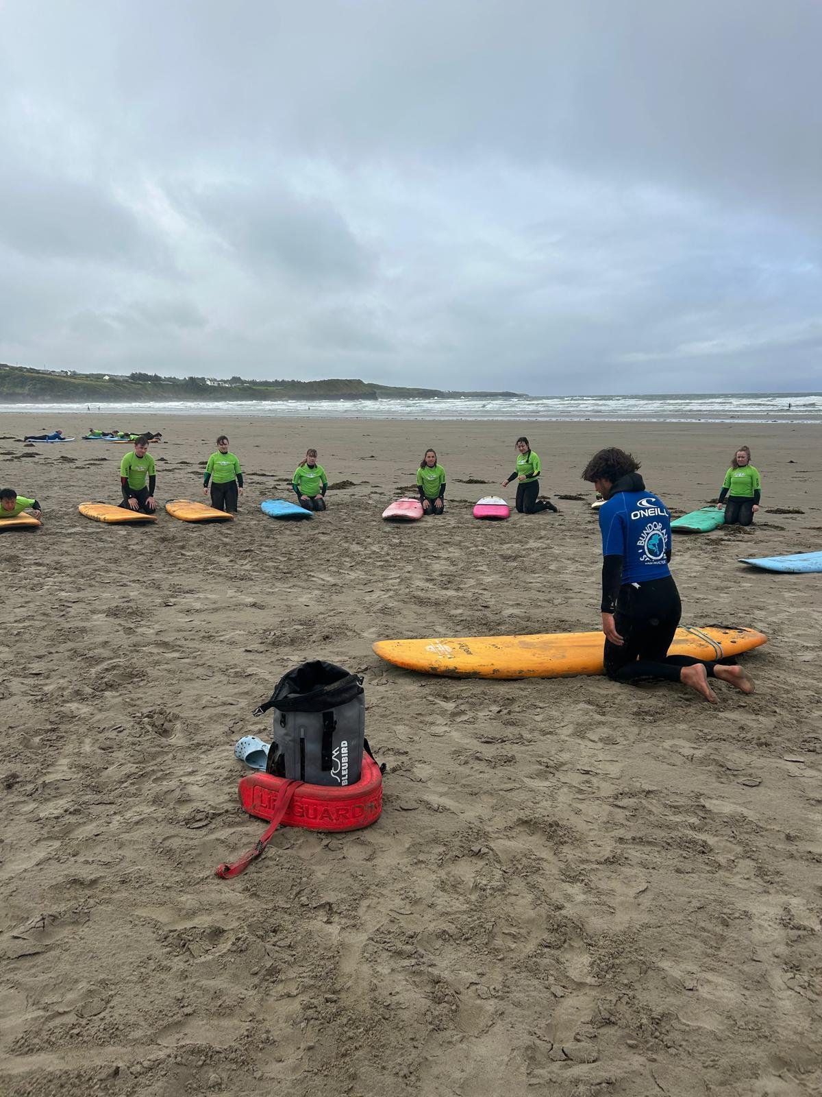 Group of people in green shirts participating in a surf lesson on a sandy beach, with surfboards lined up and a coach kneeling next to a surfboard.