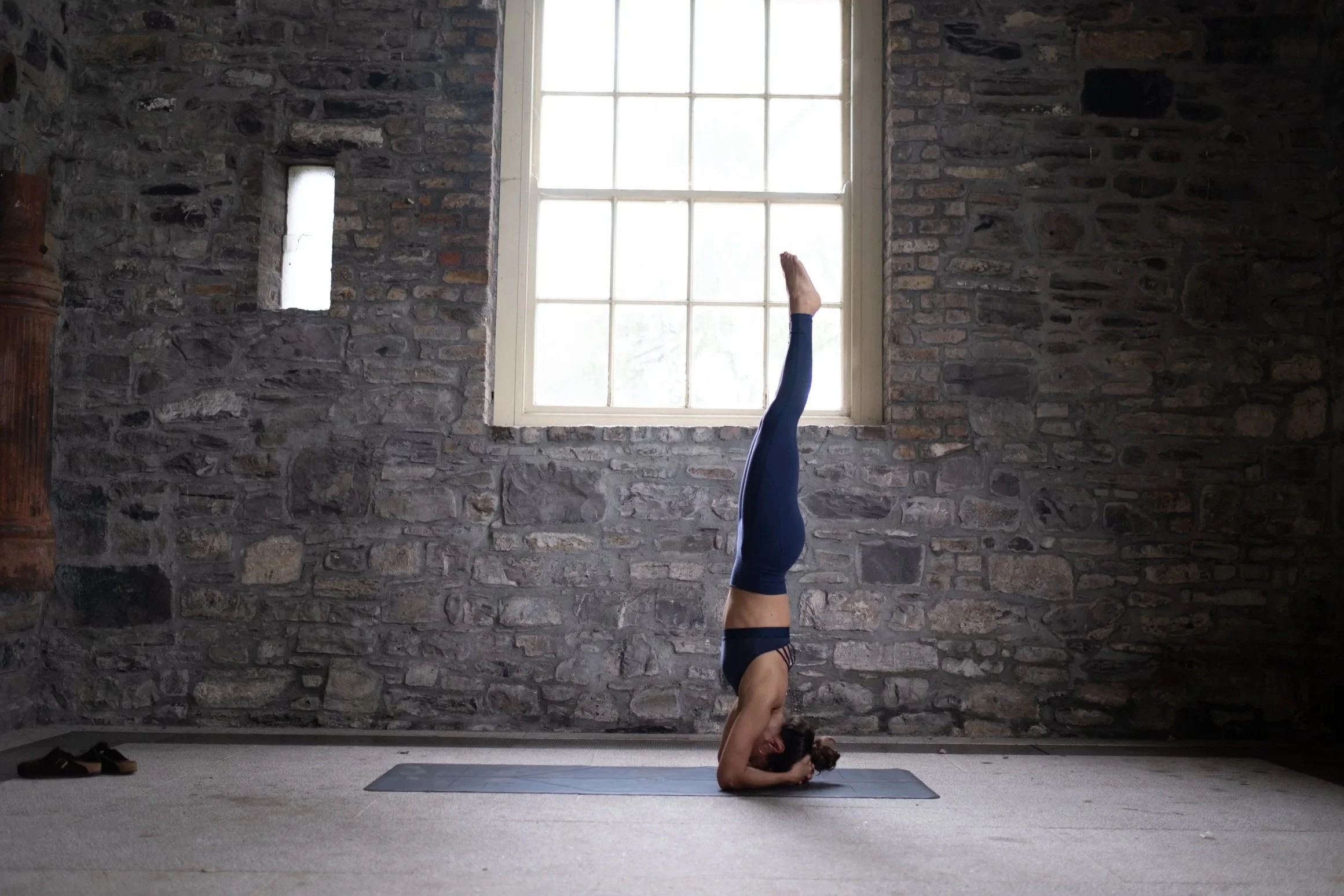 A woman practicing yoga indoors in a headstand position on a yoga mat in front of a large window with brick walls.
