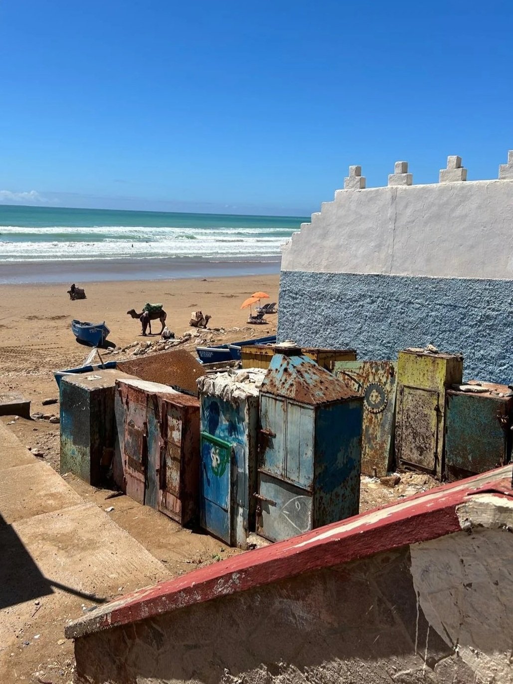 A beach with colorful, weathered metal storage boxes, a white wall with decorative steps, sandy shores, boats in the distance, and waves crashing on the shore under a bright blue sky.