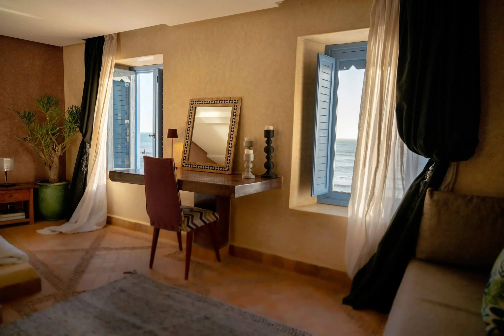 Interior of a cozy room with open blue-shuttered windows overlooking the ocean, a wooden desk with candles and a mirror, a red chair with a zigzag pattern, a potted plant, and dark and light curtains.