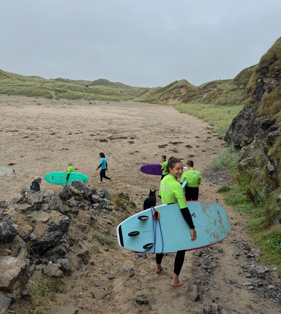 Group of people, some carrying surfboards, walking on a sandy beach with grassy dunes and rocky terrain.
