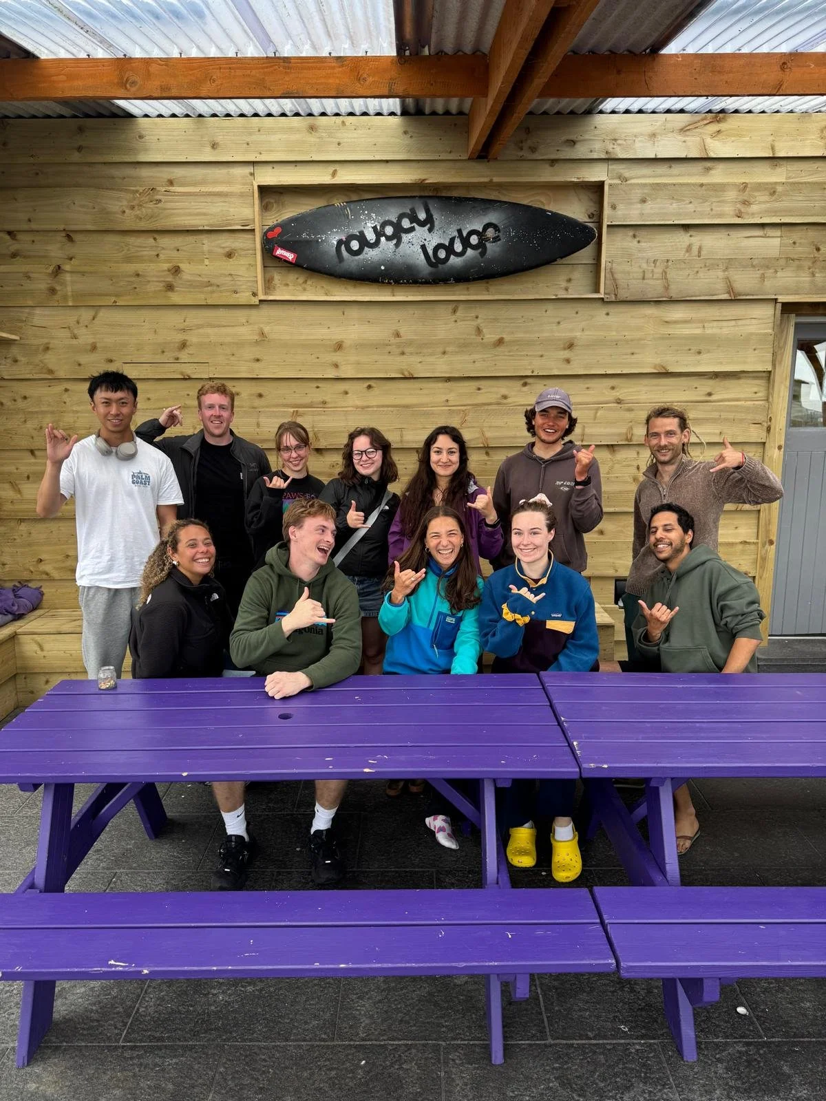 Group of ten people smiling and making hand gestures in front of a wooden wall with a surfboard that says 'Rugby Lodge' mounted above them, and purple picnic tables in the foreground.