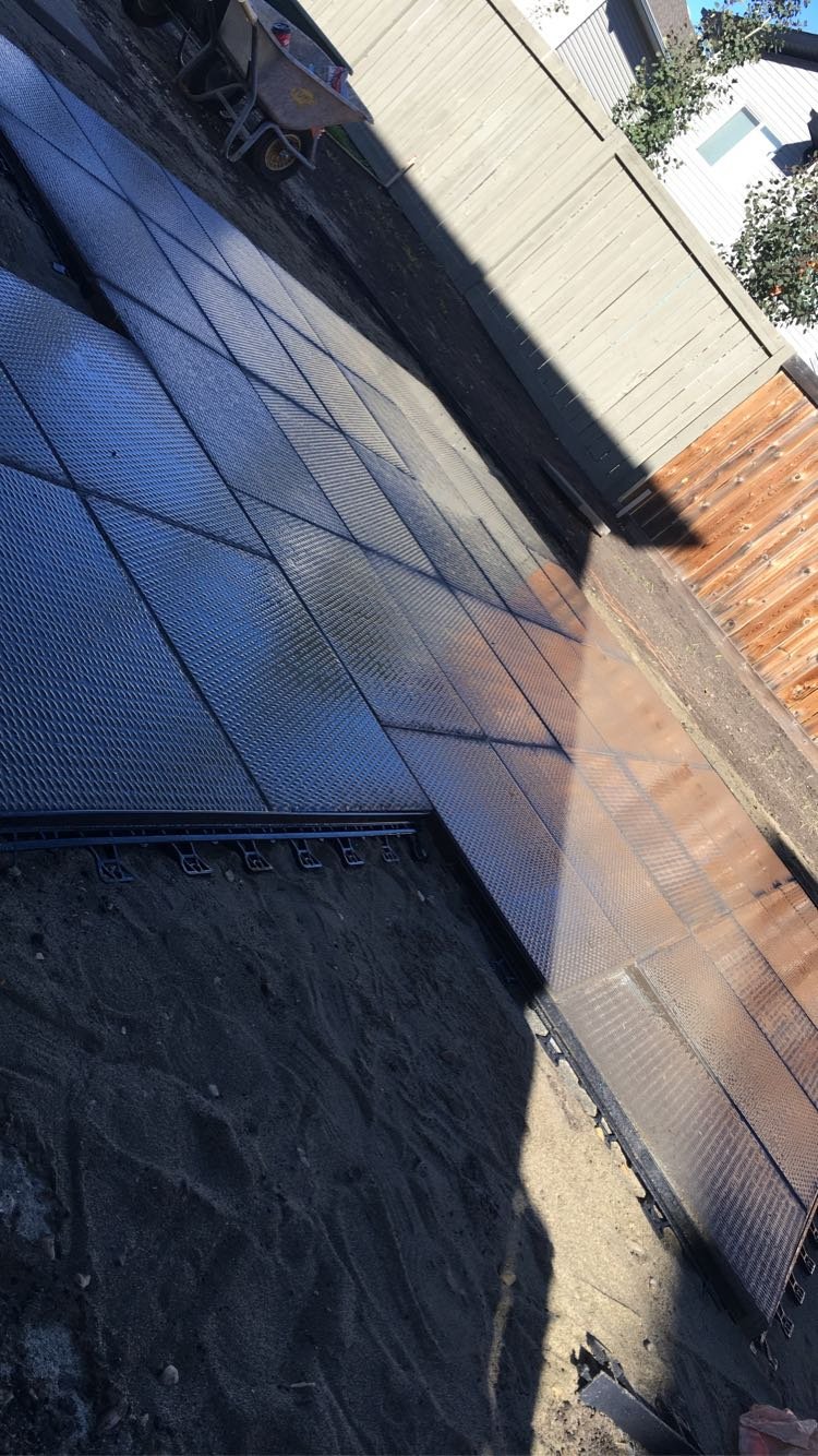 A backyard with a newly installed textured metal-plate patio or pathway, adjacent to a dirt area, with a wooden fence and some trees in the background. There is also a wheelbarrow in the upper left corner.