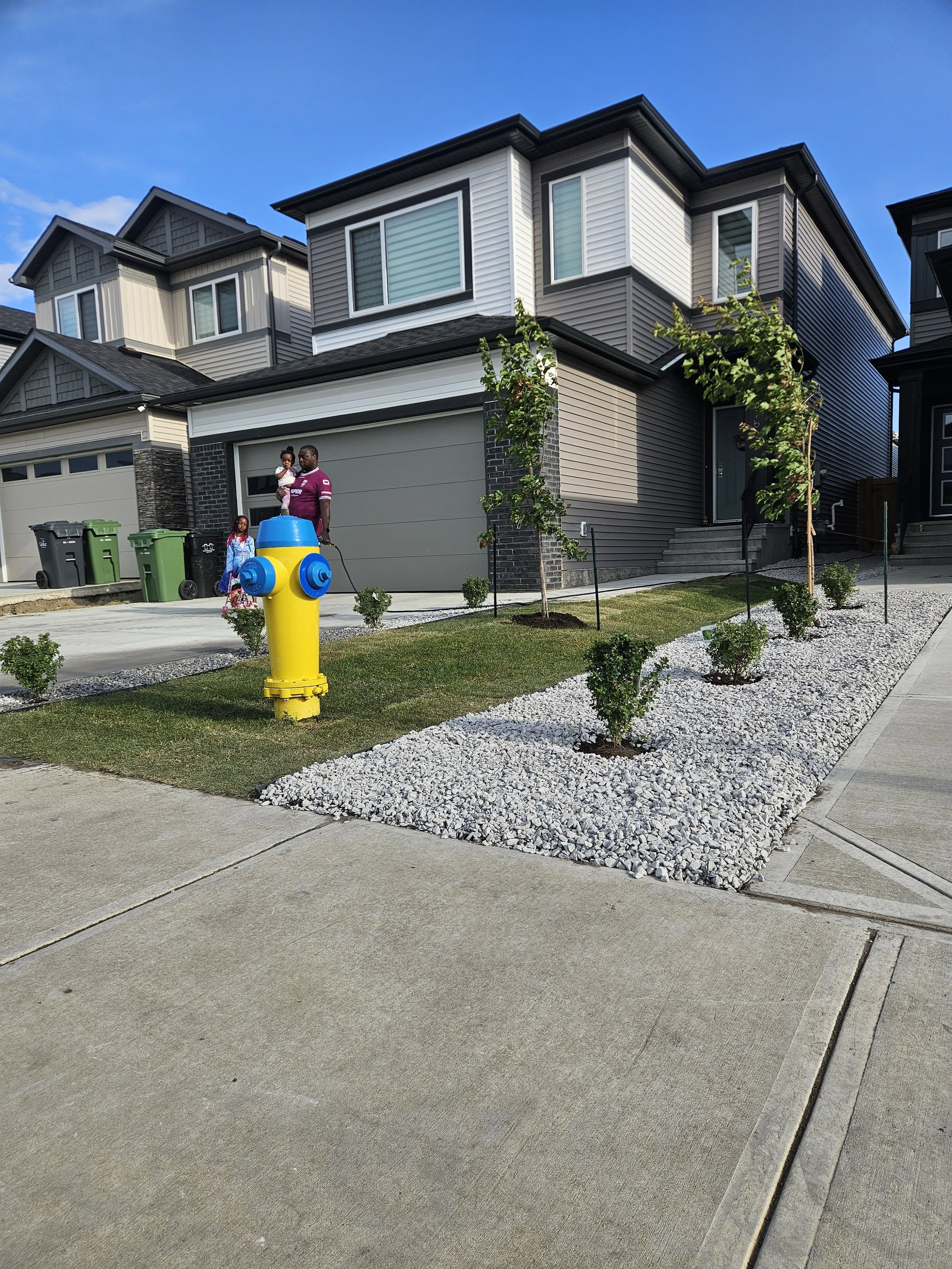 A family with two children standing outside a modern townhouse with a landscaped front yard, trees, and a yellow fire hydrant.