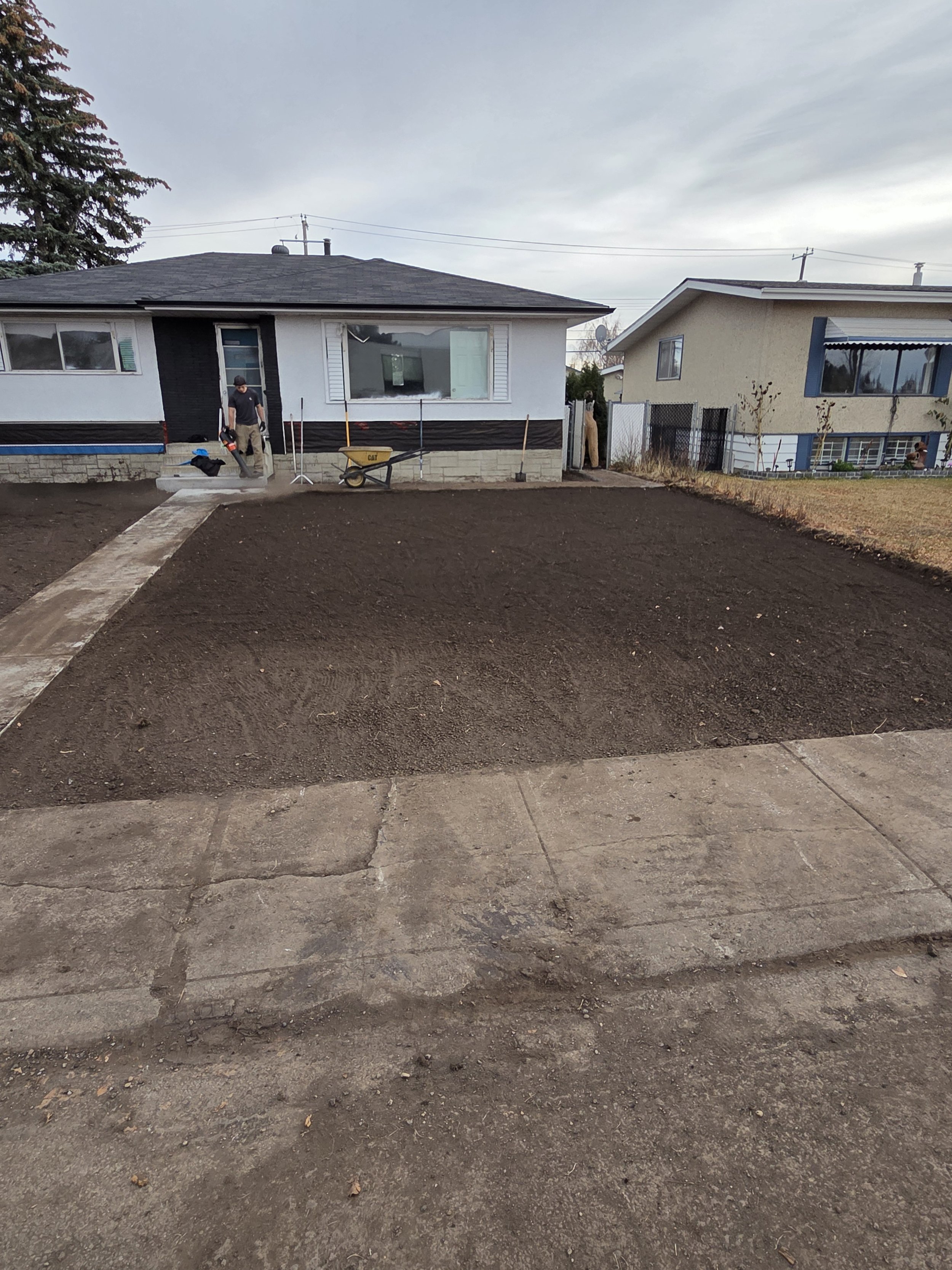 A backyard yard in front of a house with a woman and two children visible. The yard has a sidewalk on the front edge, and a flower bed area with dark soil. The house behind has a large window and a man is seen near the steps.