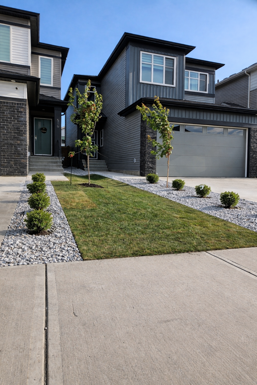 Front yard of a modern two-story house with a gray garage door, small trees, and neatly manicured lawn bordered by white rocks, with neighboring houses visible in the background.