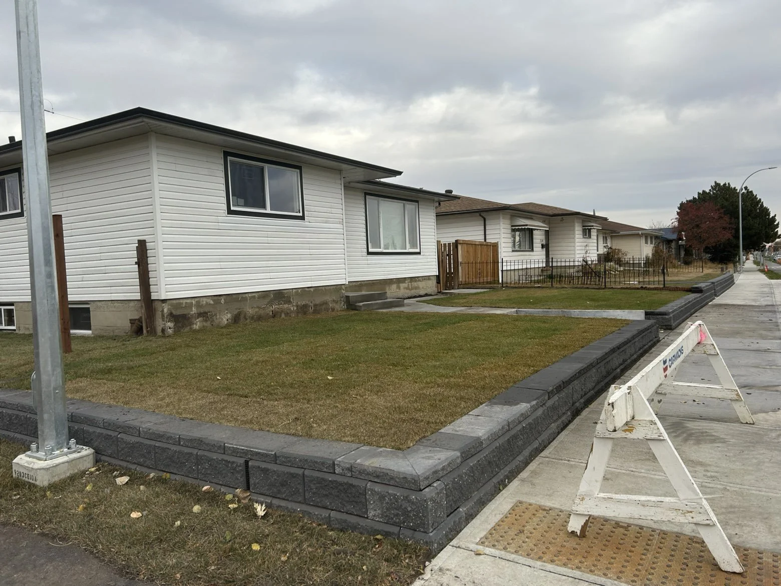 Sidewalk and new black curb stones bordering a grass lawn; white house with dark window frames; construction barriers in the foreground; cloudy sky.