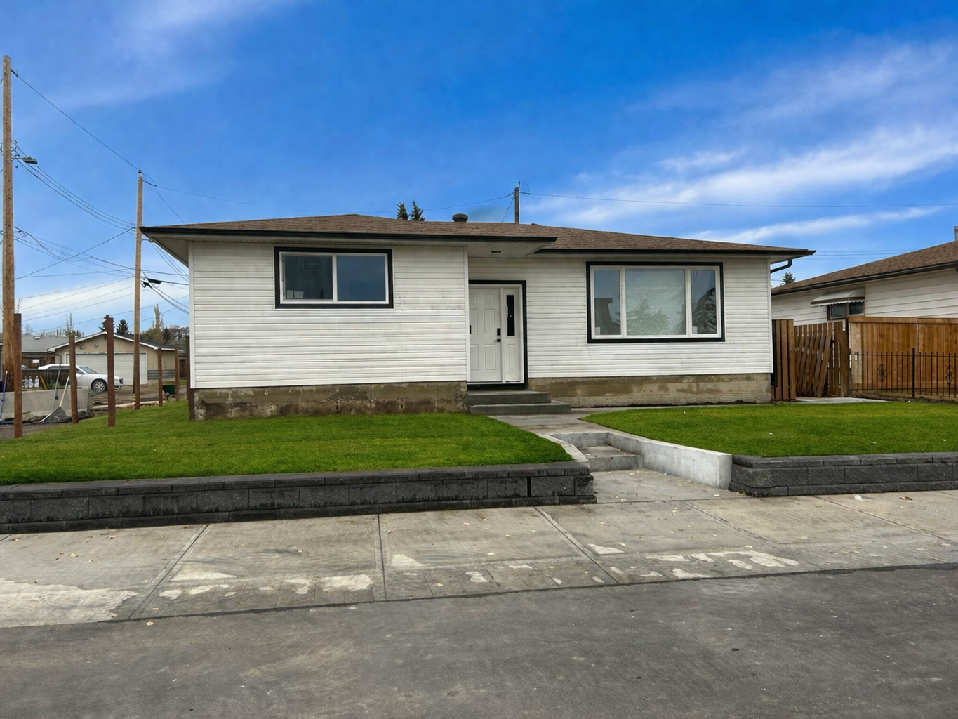 A white single-story house with black trim, a front door with a small staircase, and two large windows, surrounded by a green lawn and a concrete sidewalk in a suburban neighborhood.