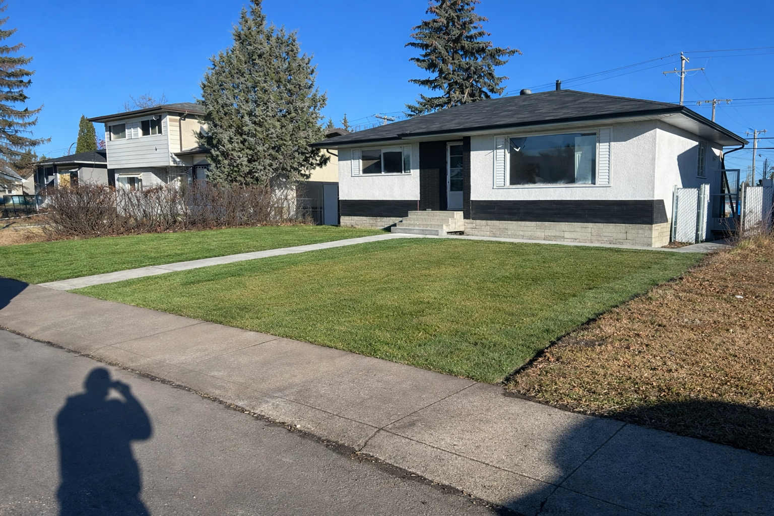 A single-story house with a black and white exterior, front step, large window, and front lawn with a walkway. Nearby trees and neighboring houses are visible against a clear blue sky.