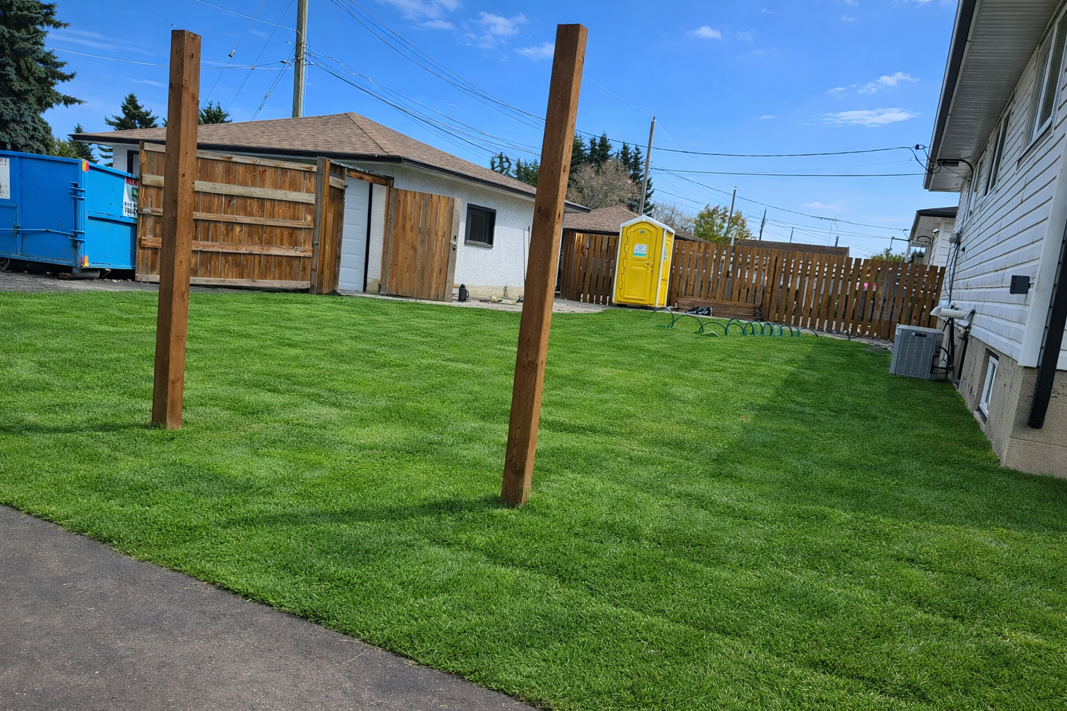 A backyard with a well-maintained green lawn, a wooden fence, and a small white building with a door. There are four wooden posts in the grass, a yellow portable toilet, a blue dumpster, and a garden hose on the ground. The edge of the house with an air conditioning unit is visible on the right.