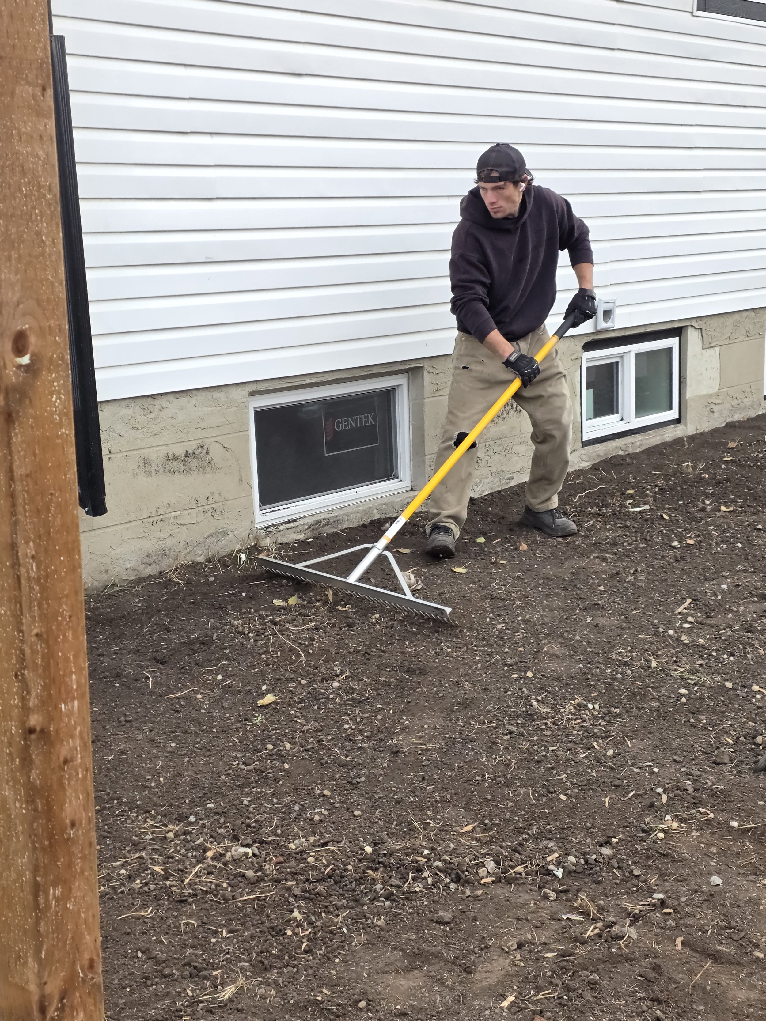 Man in black hoodie, khaki pants, and black cap using a rake to level soil outside a house with white siding and small basement windows.