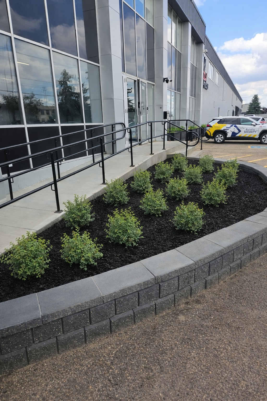 Exterior view of a modern commercial building with a sloped concrete wheelchair ramp and railing leading to glass doors. In front, there is a landscaped section with small green bushes bordered by gray bricks. To the right, there is a parking lot wit