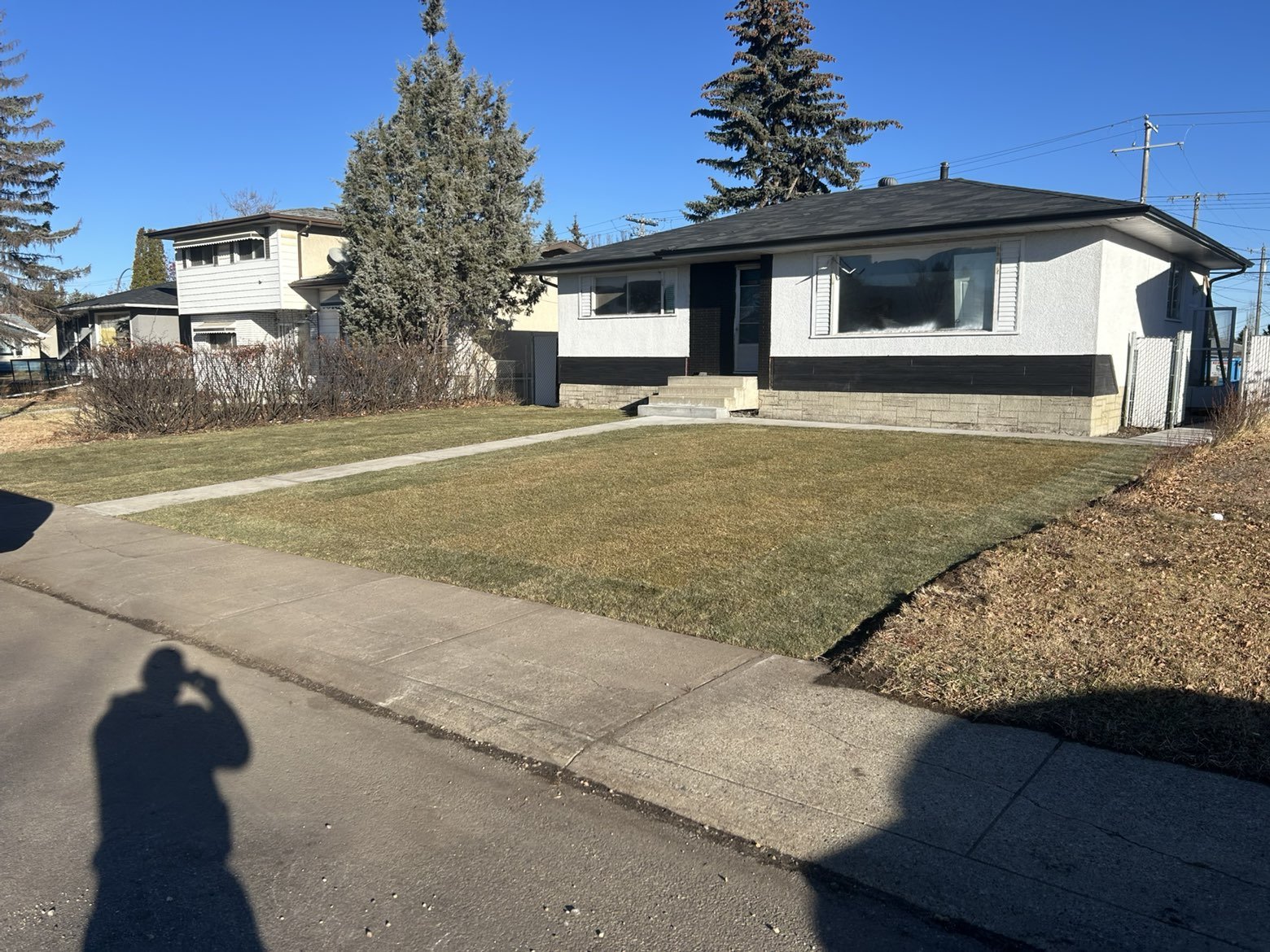 A single-story white house with black trim and steps leading to the front door, surrounded by a lawn and sidewalk, with neighboring houses and tall trees in the background under a clear blue sky.