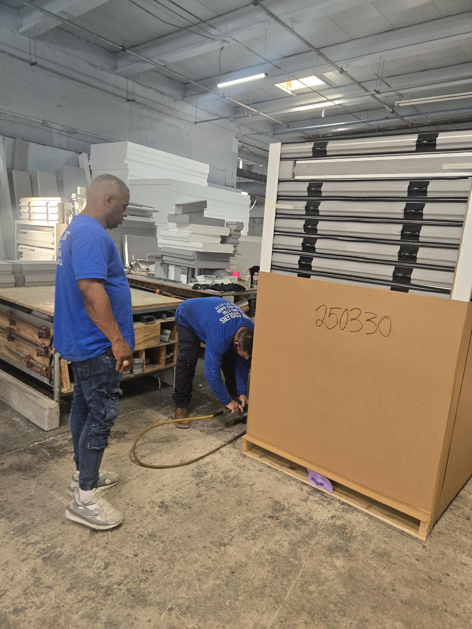 Two workers in blue shirts are handling a large cardboard box labeled '250330' in a warehouse, with one standing and the other bent over working on the box.