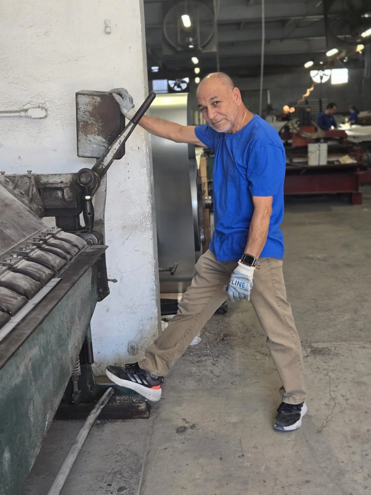 An older man in a blue shirt, beige pants, sneakers, and a smartwatch presses his foot against a large industrial machine in a workshop or factory setting.