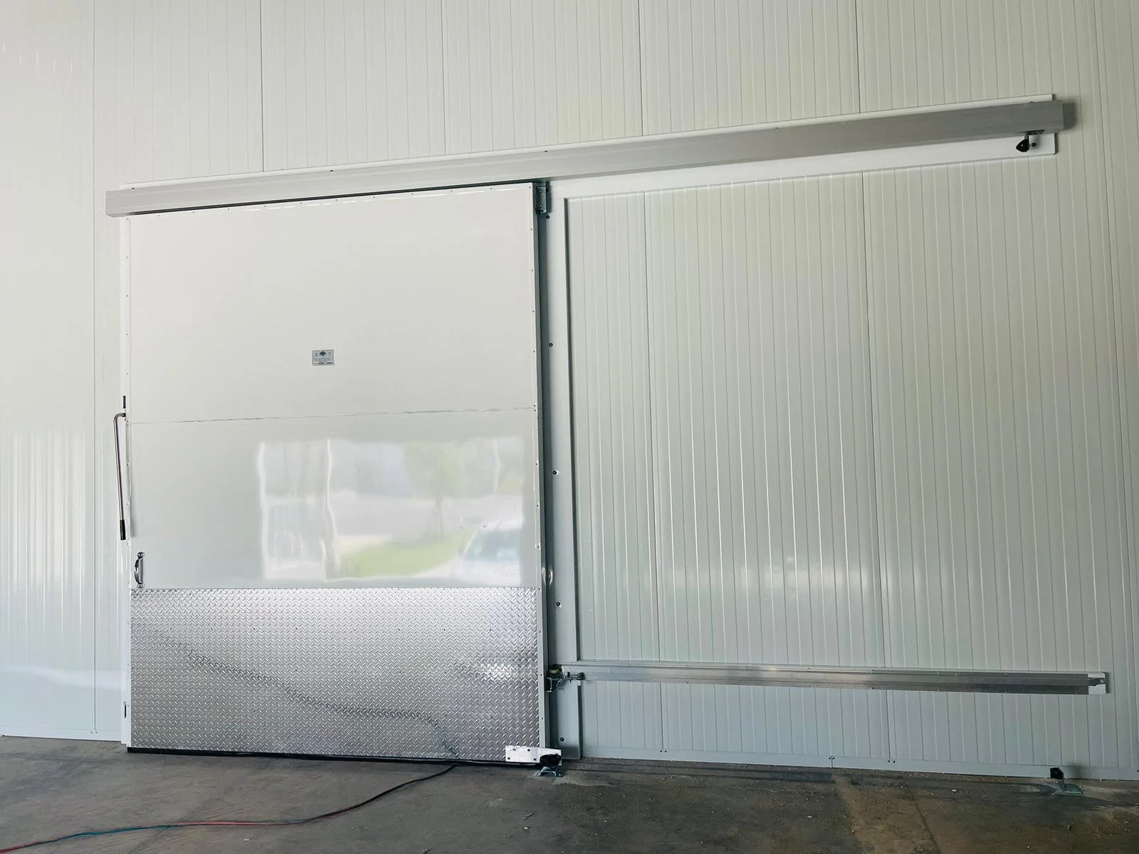 White industrial sliding door with diamond plate at the bottom, installed on a metal paneled wall in a warehouse or garage.