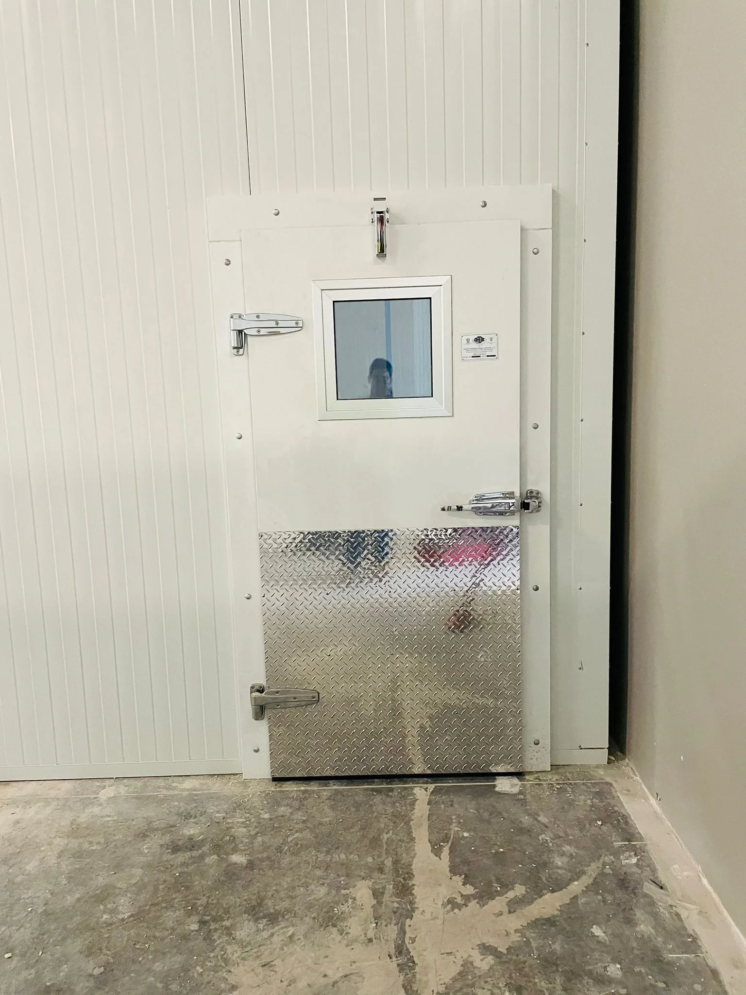 A metal door with a small window, set in a wall with white vertical paneling. The door has metallic handles and locks, and a silver diamond plate panel at the bottom.