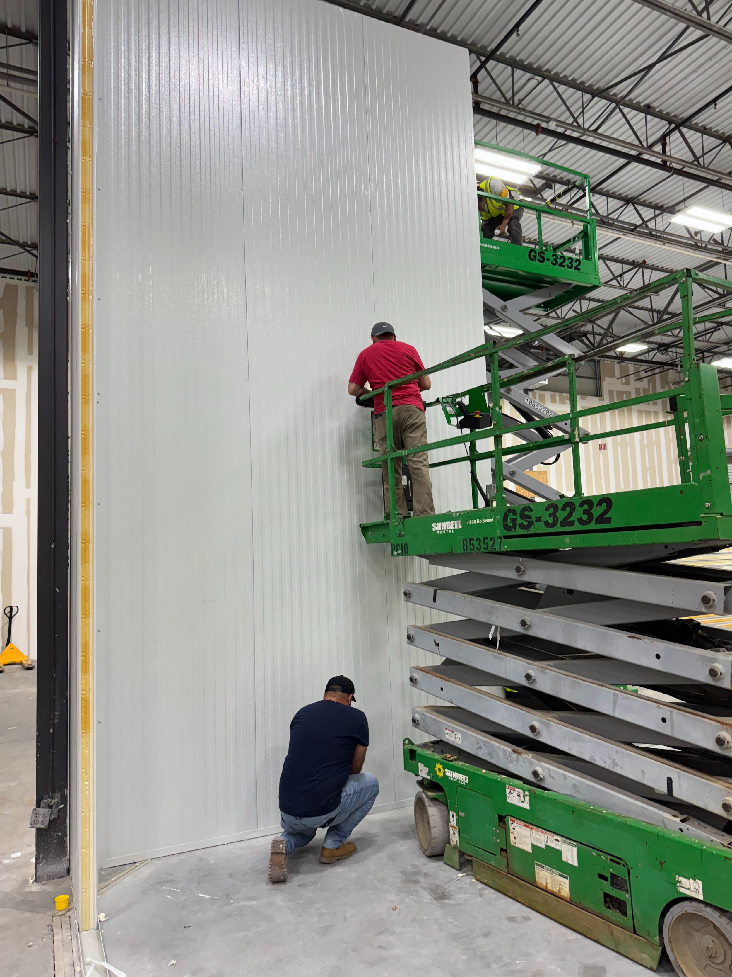 Two workers on green scissor lifts installing or working on a white corrugated metal wall inside a large industrial building.