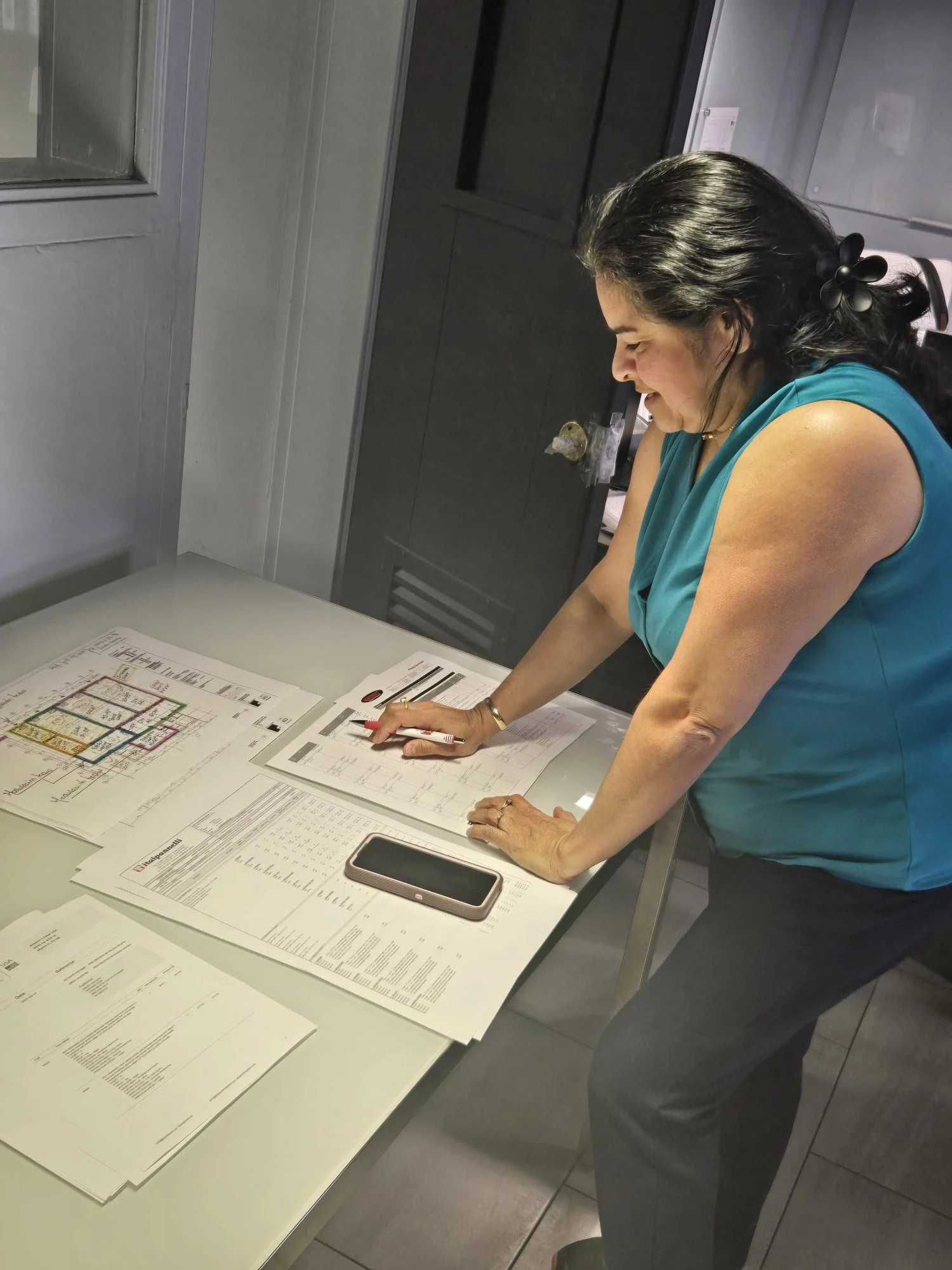 Woman in a teal sleeveless top working at a table with papers and a smartphone, leaning forward and reading documents, in a gray room with a small window.