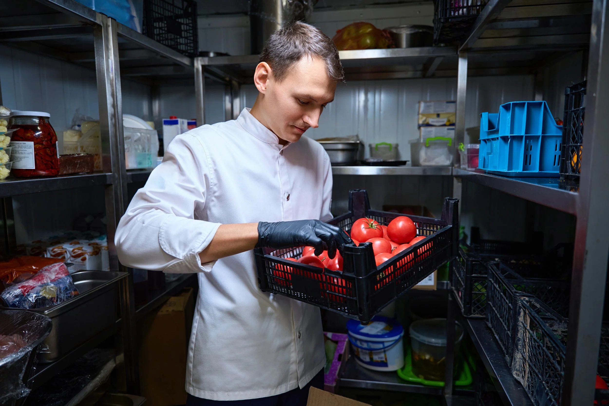 A man wearing a white chef's coat and black gloves inspecting a crate of red tomatoes in a storage room.