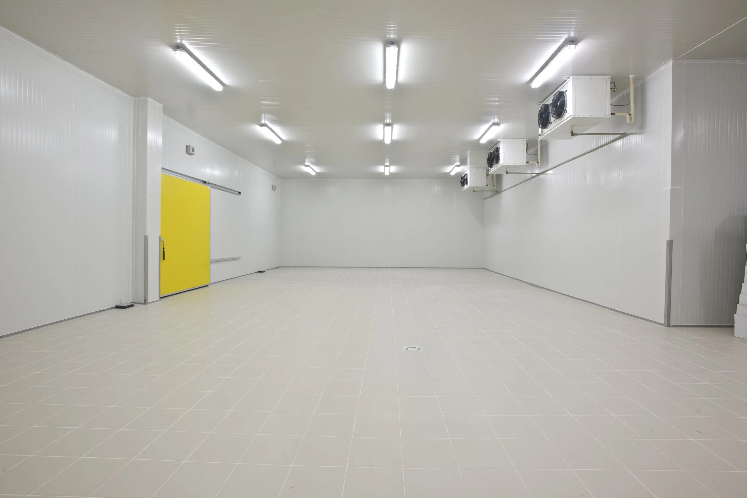 Empty industrial or commercial room with white walls, white ceiling, beige tiled floor, fluorescent lighting, a yellow door, and cooling units on the wall.