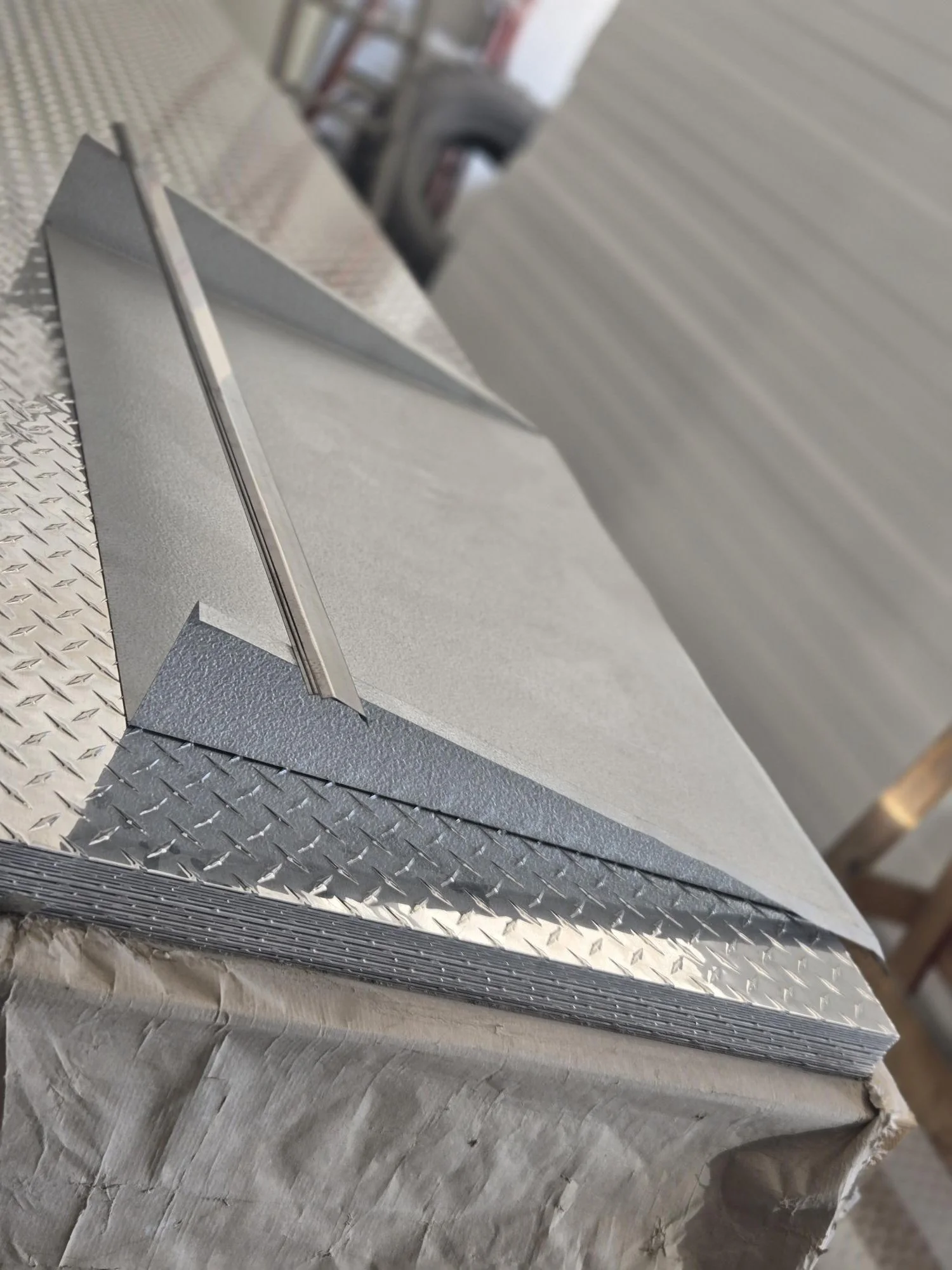 Close-up of a metal L-shaped piece resting on a diamond plate workbench, with an unfinished wall in the background.