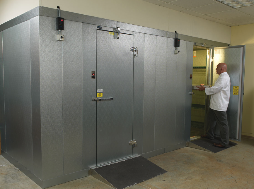 A man in a white shirt standing inside an industrial walk-in refrigerator or cold storage room, holding a clipboard.