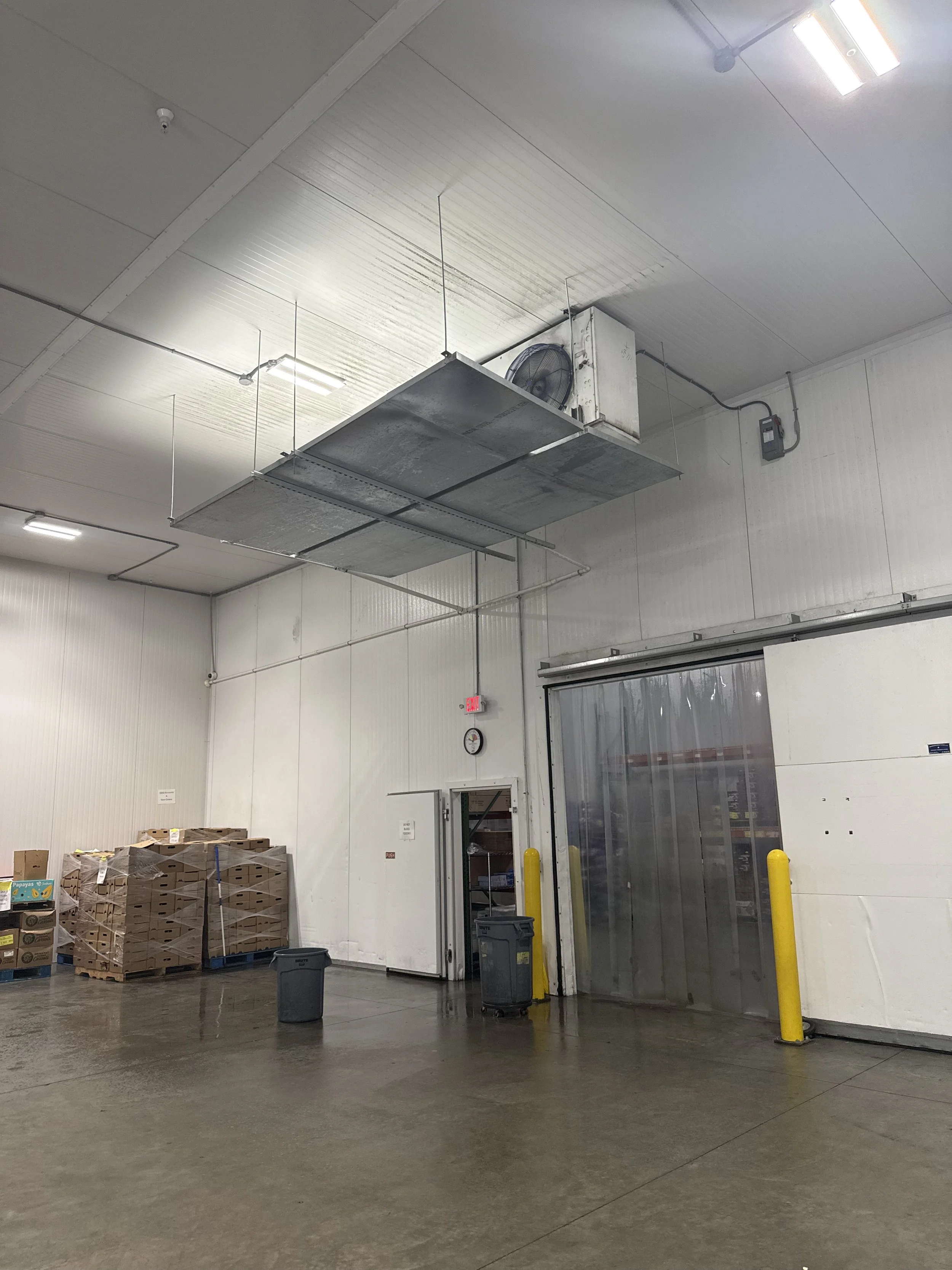 Industrial warehouse with a high ceiling, large metal air ventilation units, stacked cardboard boxes on pallets, trash cans, and a sliding door.