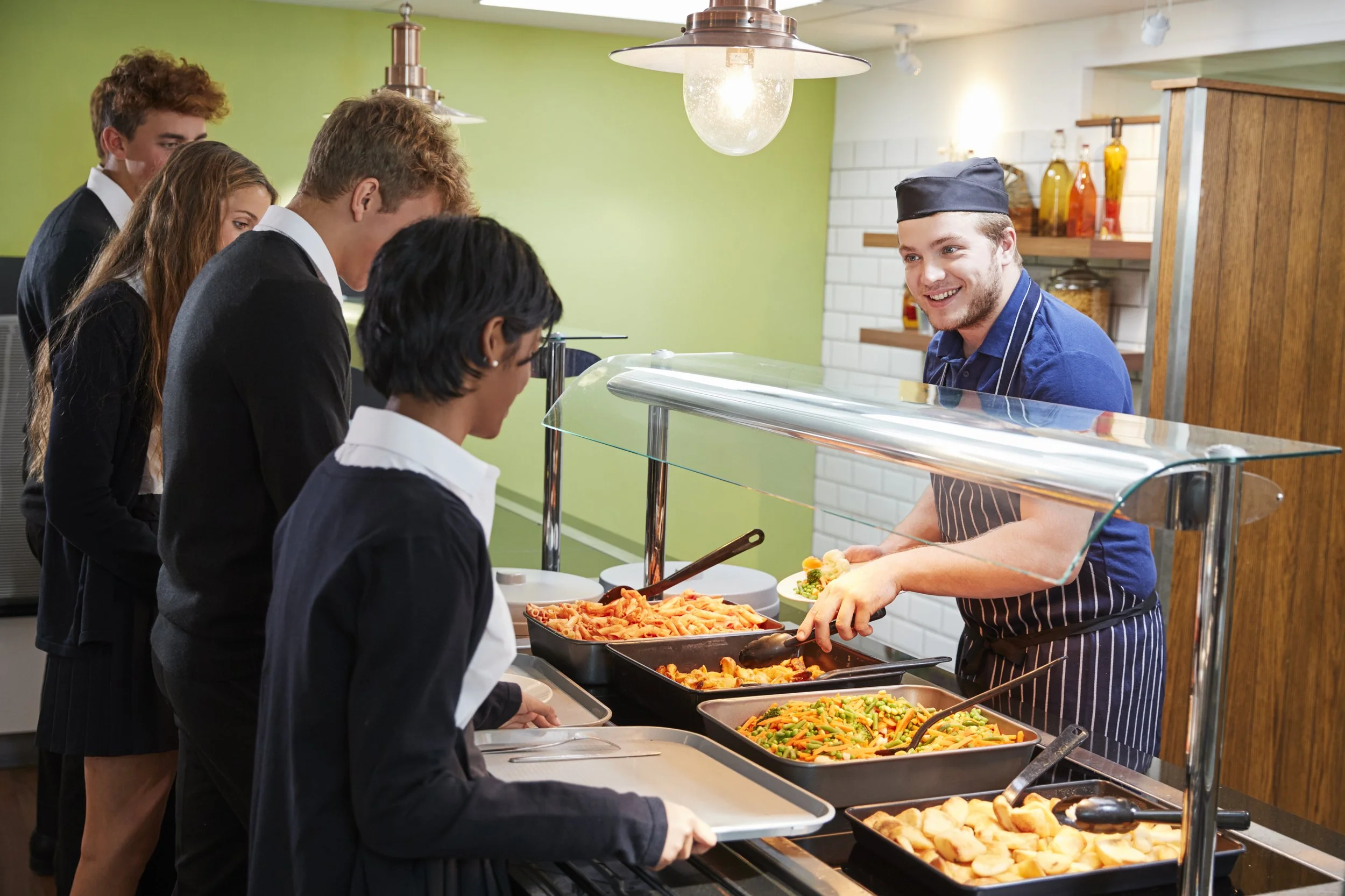 A group of students in school uniforms lining up at a cafeteria with a smiling worker serving food.